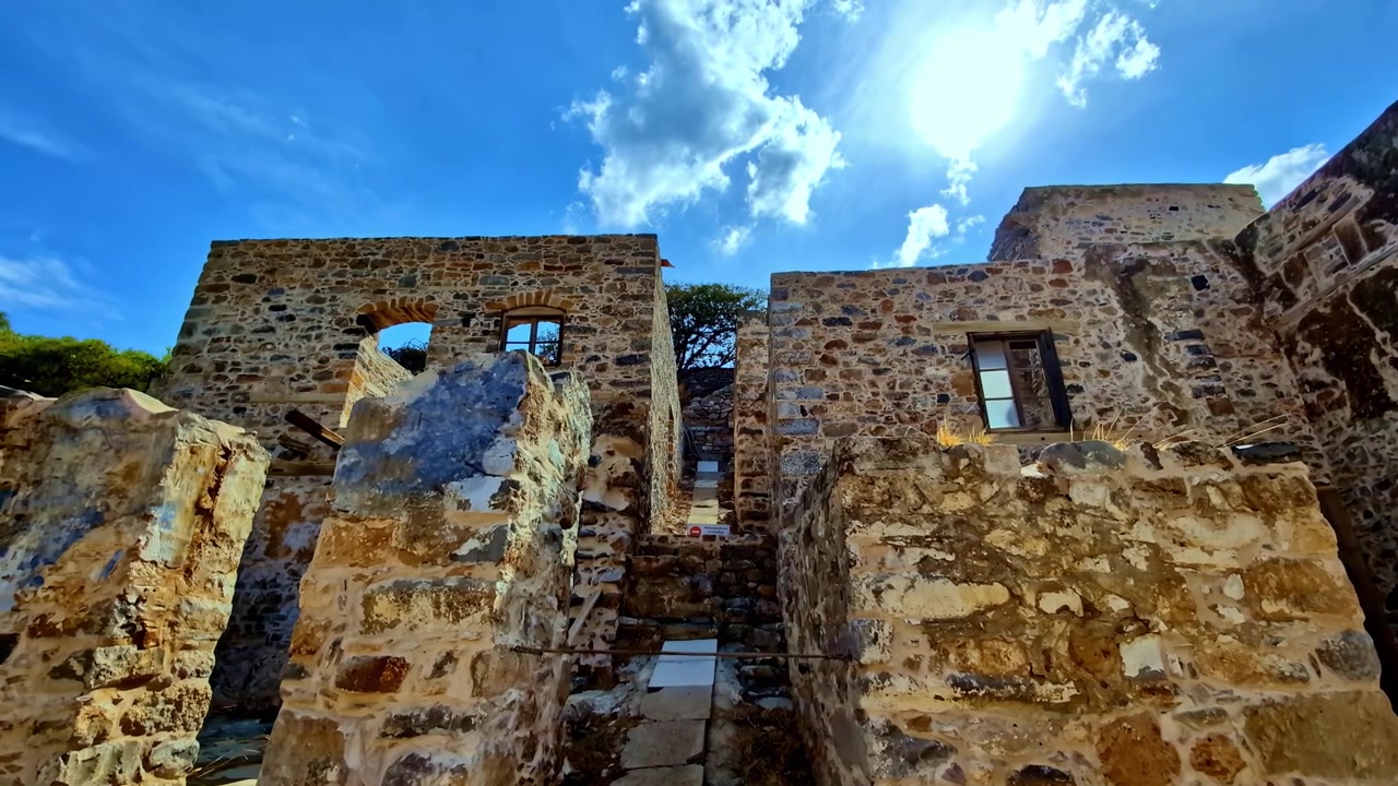 Le mura in pietra consumate della fortezza di Spinalonga si ergono sotto un sole splendente di mezzogiorno mentre la telecamera riprende dal cortile in rovina.