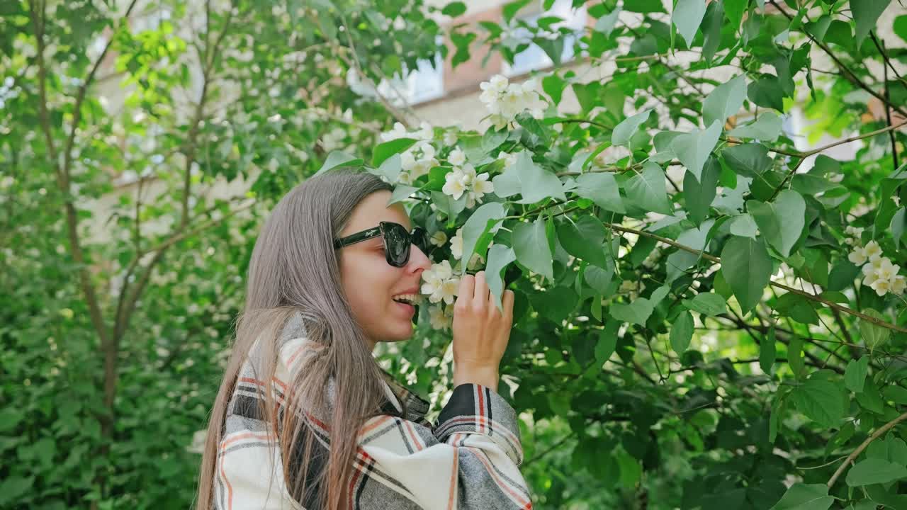 Young woman gently smells blooming jasmine flowers, green courtyard slow motion