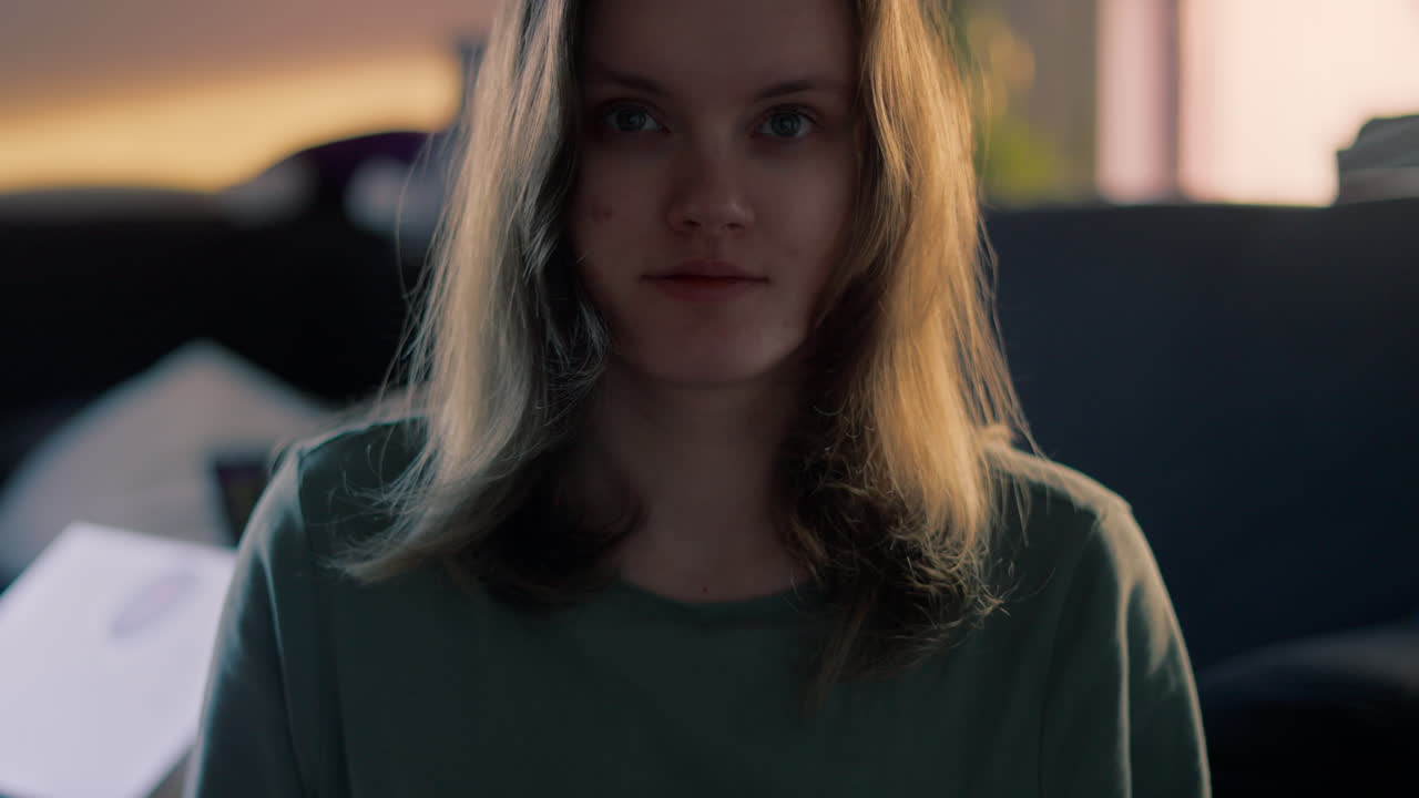 Young white woman staring intensly straight into the camera with wide eyes open and subtle smile during a golden hour at sunrise in her living room at home in slow motion