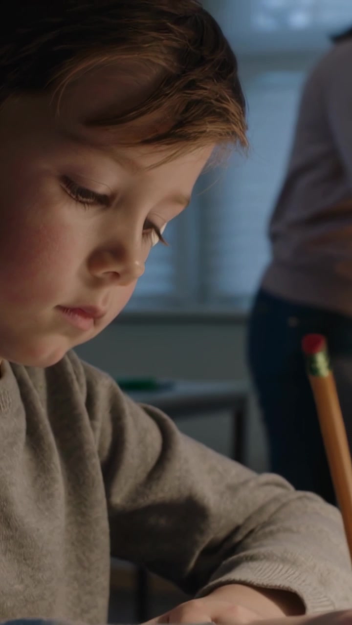 Video verticale: Matita che tocca la carta, bambino con maglietta grigia che scrive al tavolo vicino alla finestra, con un genitore che osserva.