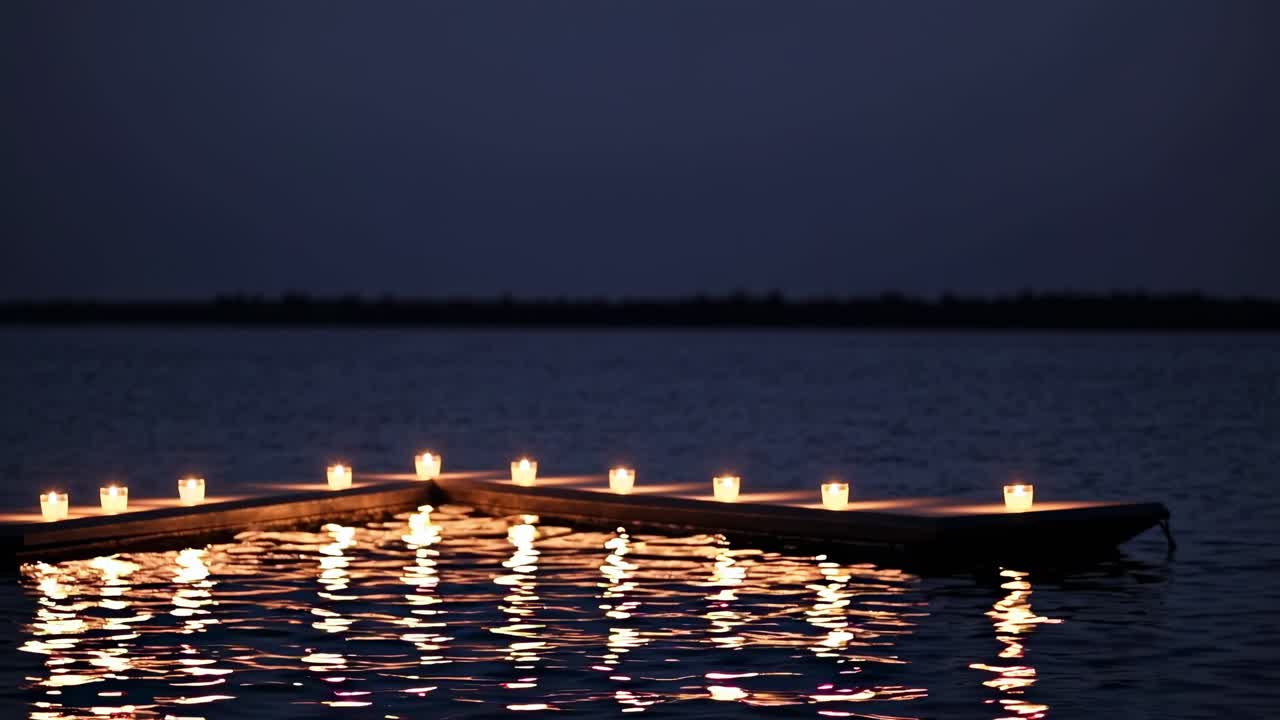Several lit candles are placed on a wooden dock floating on calm water, creating a serene and romantic atmosphere during twilight, with the soft glow reflecting on the water's surface