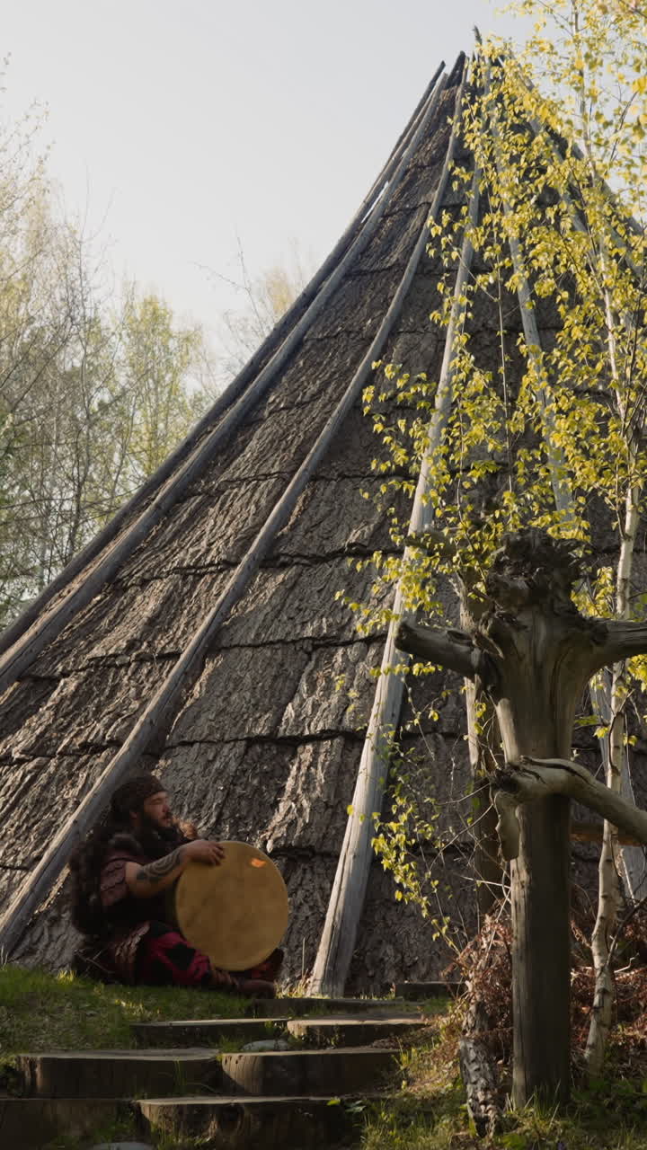 Obese man in ethnic clothes holds shamanic drum sitting near old ail building. Decorations for folk culture festival. Life in ancient village