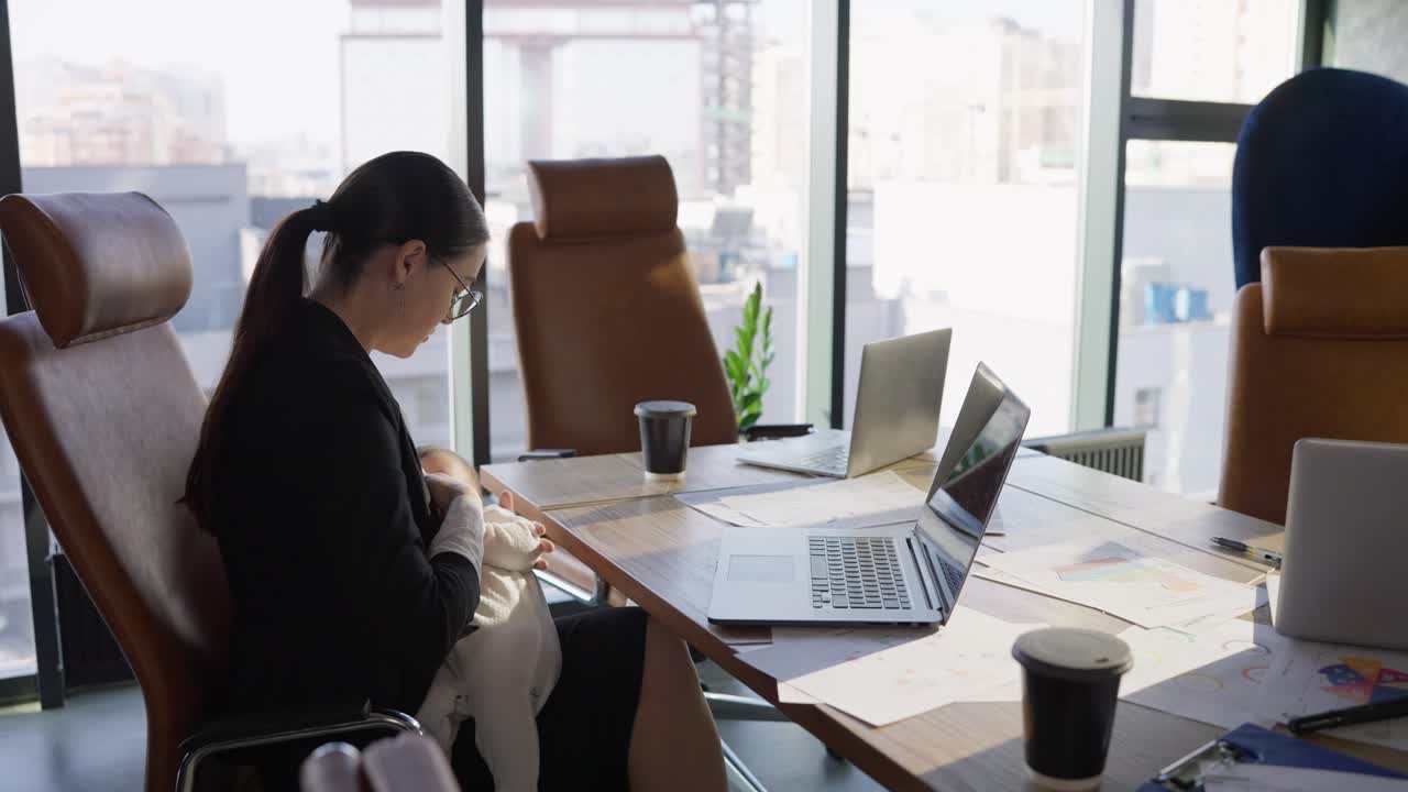 Confident brunette businesswoman in business clothes sits at an office desk in front of a laptop and breastfeeds her nursing baby In the office