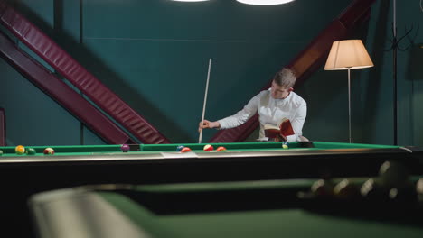 young player in white shirt reads red book with focused expression while holding cue stick near green pool table with scattered billiard balls. soft lighting from overhead lamp