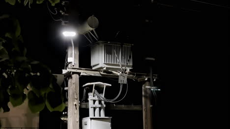 Closeup-shot-at-night,-tall-white-transformer-electricity-pole-next-to-city-tree