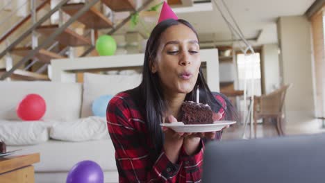 mixed race woman using laptop having birthday video chat holding a cake blowing candle