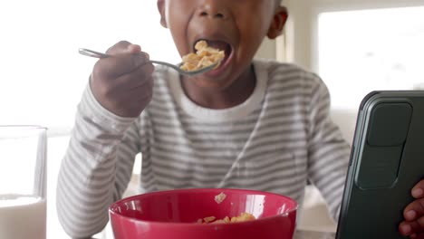 happy african american boy having breakfast and using tablet in kitchen, slow motion