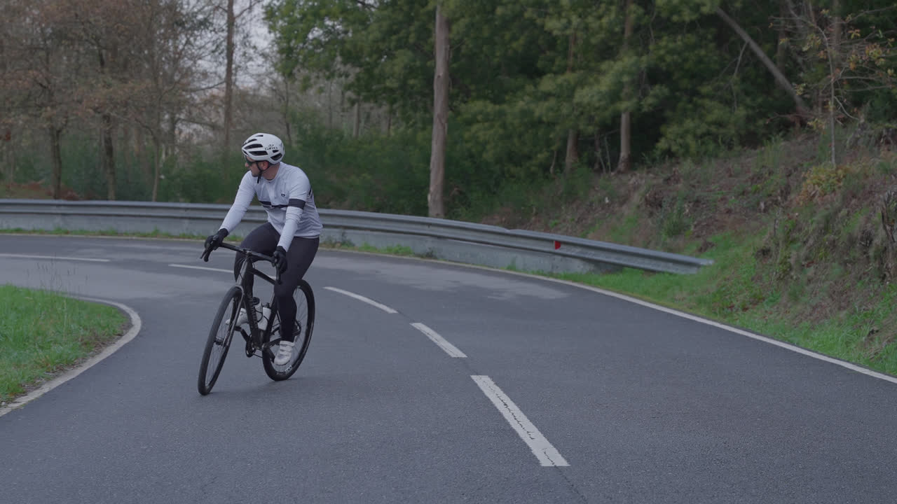 Uomo in bicicletta in salita su strada asfaltata con guardrail e alberi.