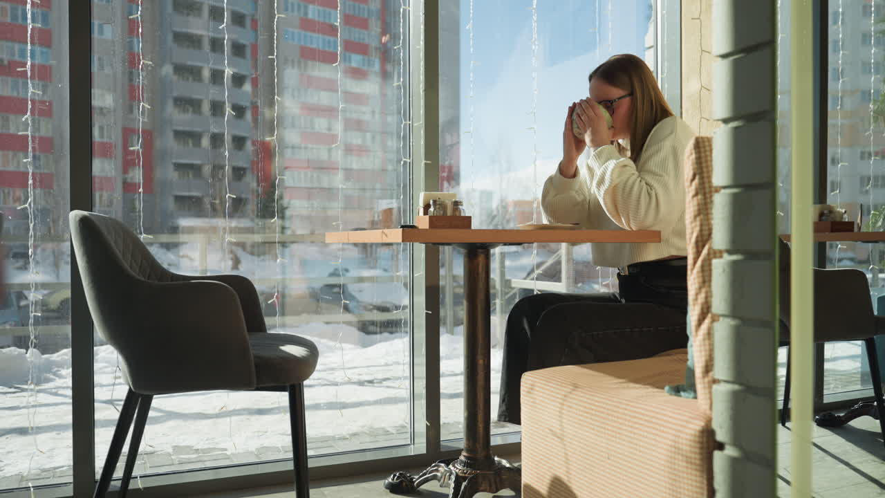 Woman seated at table sipping tea in cozy cafe setting with winter sunlight streaming through large windows, outside view features residential buildings, parked cars, and passing pedestrians in snow