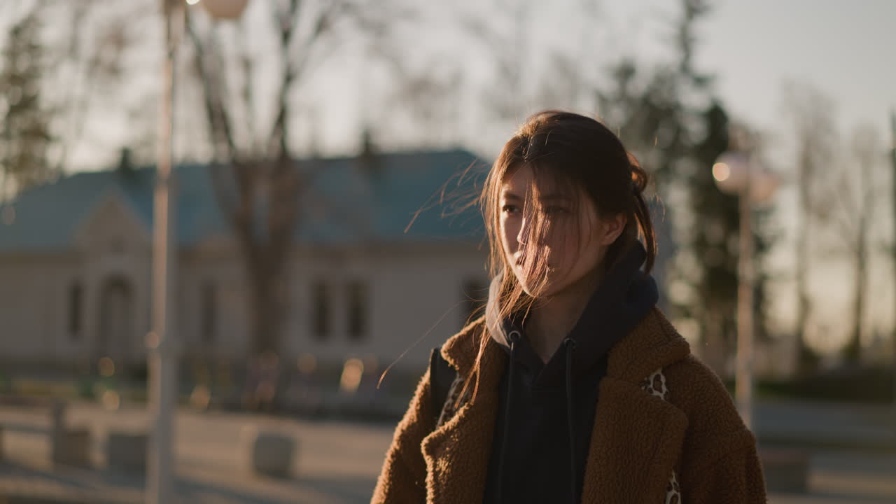 갈색 코트와 후디를 입은 소녀는 피곤하고 상하는 표정으로 희미하게 고 있습니다. 그녀의 머리카락은 바람에 부드럽게 어지고 있습니다. 도시 환경에서 조용한 반사 순간.