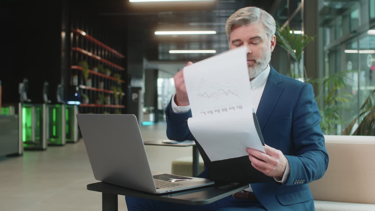 Happy male entrepreneur in suit analyzing graph documents reports and using laptop in office lobby
