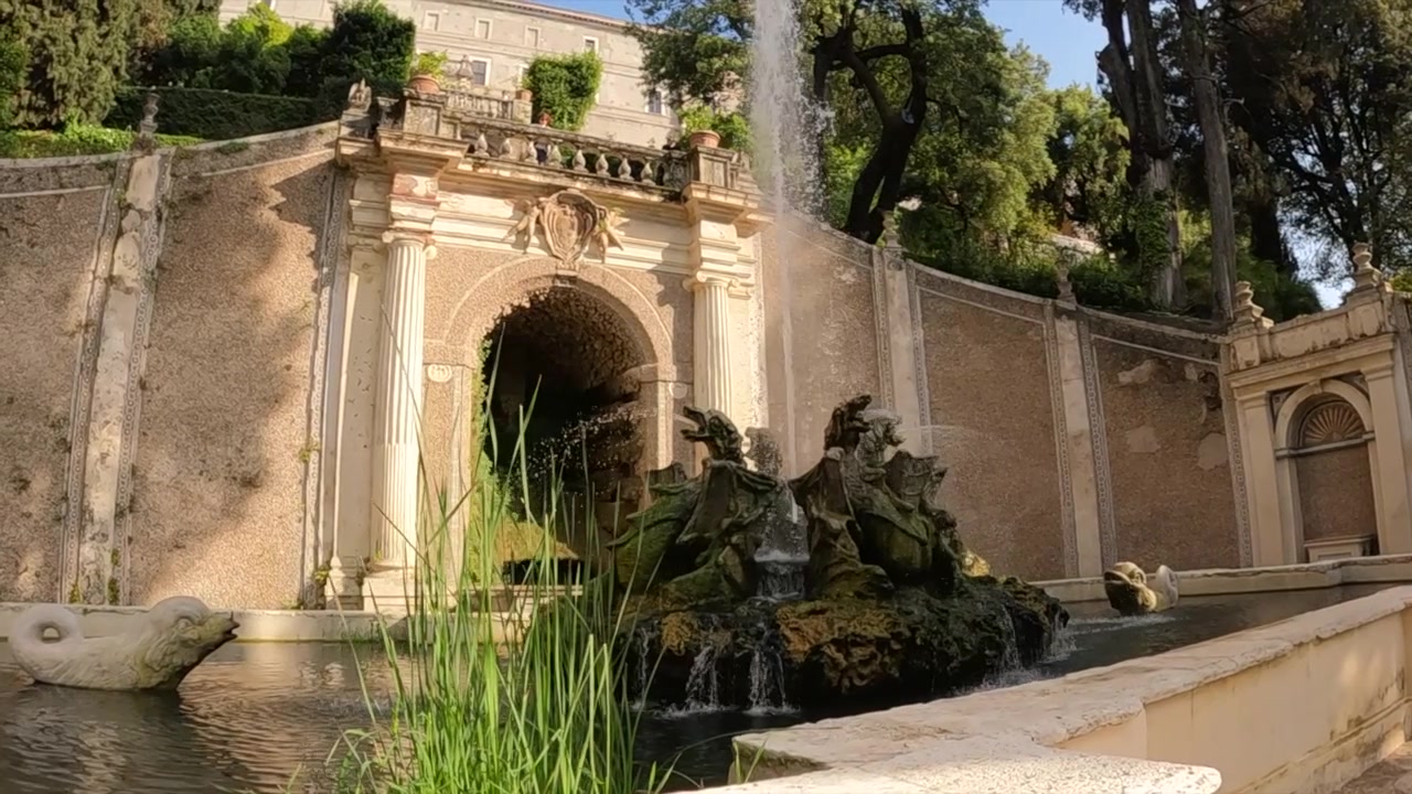 Villa d'Estes barokfontæne med skulpturerede figurer, kaskaderende vand, udsmykket stenbuegang og frodigt grønt.