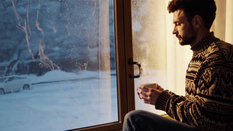 man drinking warm beverage by window on a cold winter day