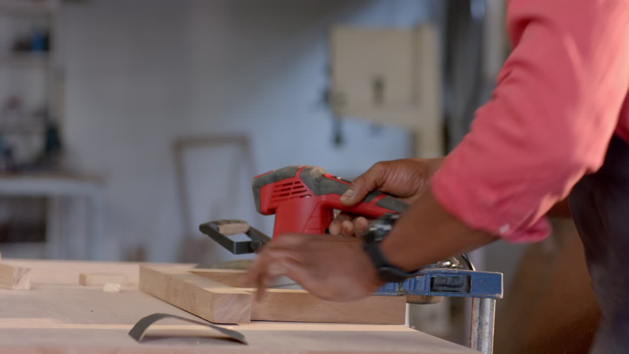 Sanding senior African American man wearing apron using sander on plank clamped to workbench