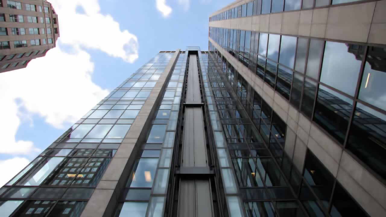 Looking upward from the ground, the view captures the towering skyscraper with sleek glass windows and a bright sky backdrop.