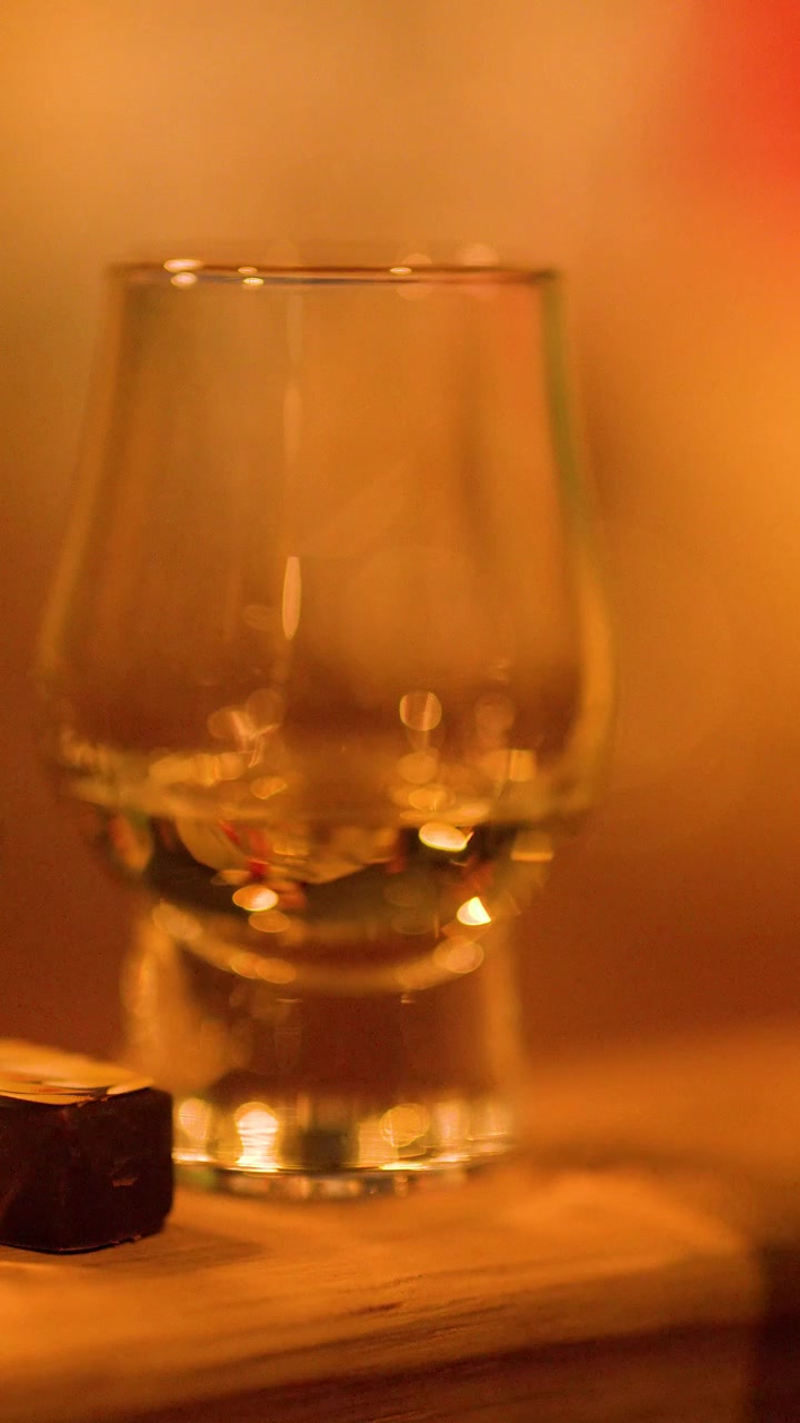 Una mano solleva un bicchiere di whisky da una superficie di legno in una calda illuminazione ambientale da bar.