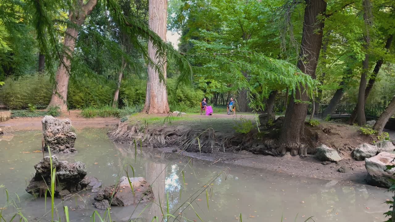 filmación de un lugar bucólico donde una pareja de hombre y mujer aparecen en una isla al lado de una secuoia sentada en una silla de madera, ella lleva una falda de un color llamativo, la vegetación es impresionante