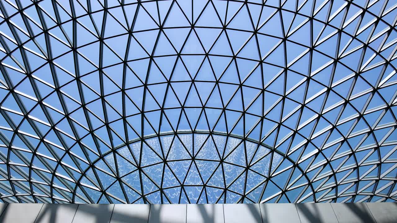 Camera tilts upward beneath a geometric glass dome, revealing a large indoor waterfall with natural daylight illuminating the architectural structure