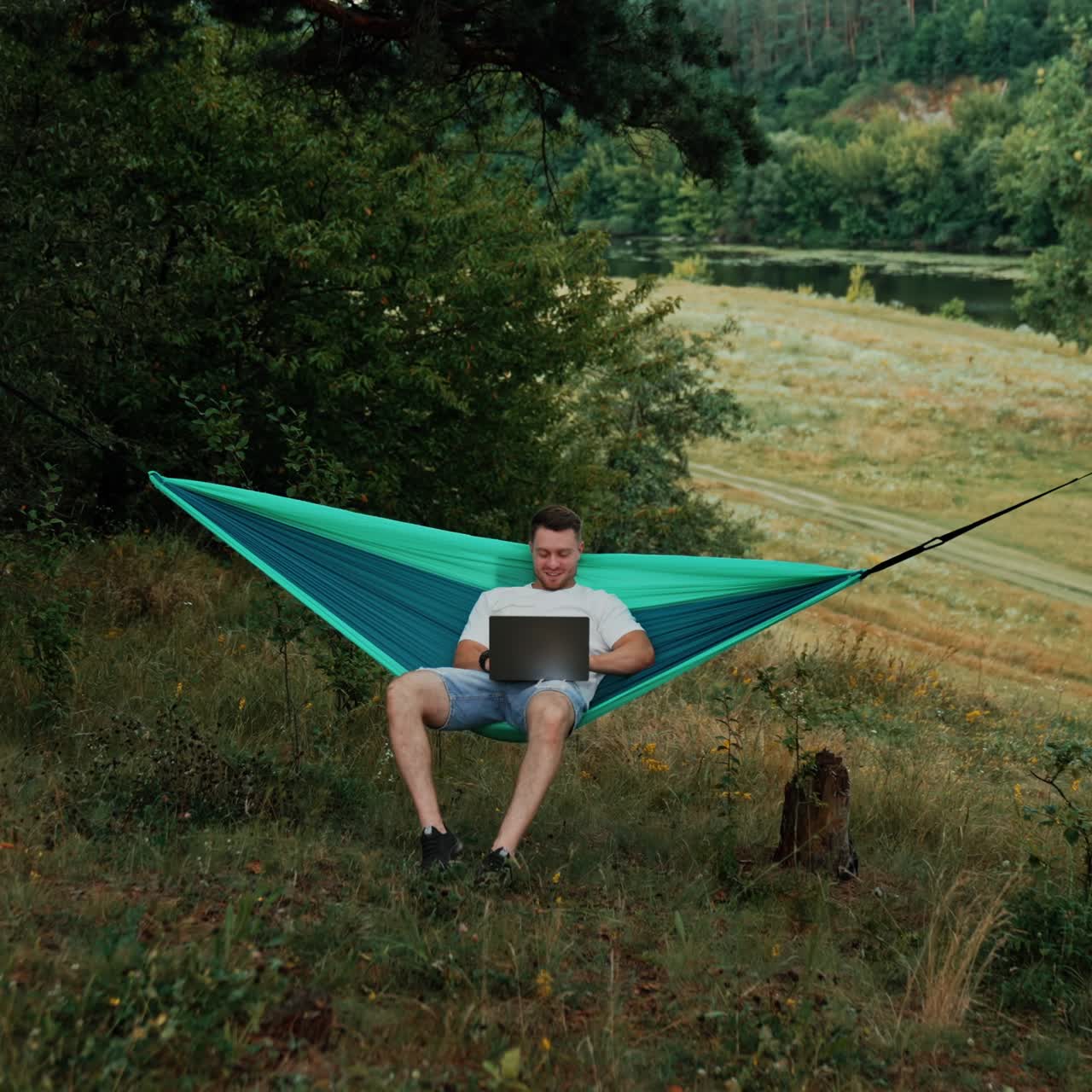 Caucasian man sits swaying in the hammock. Freelancer doing remote job on his laptop in the nature