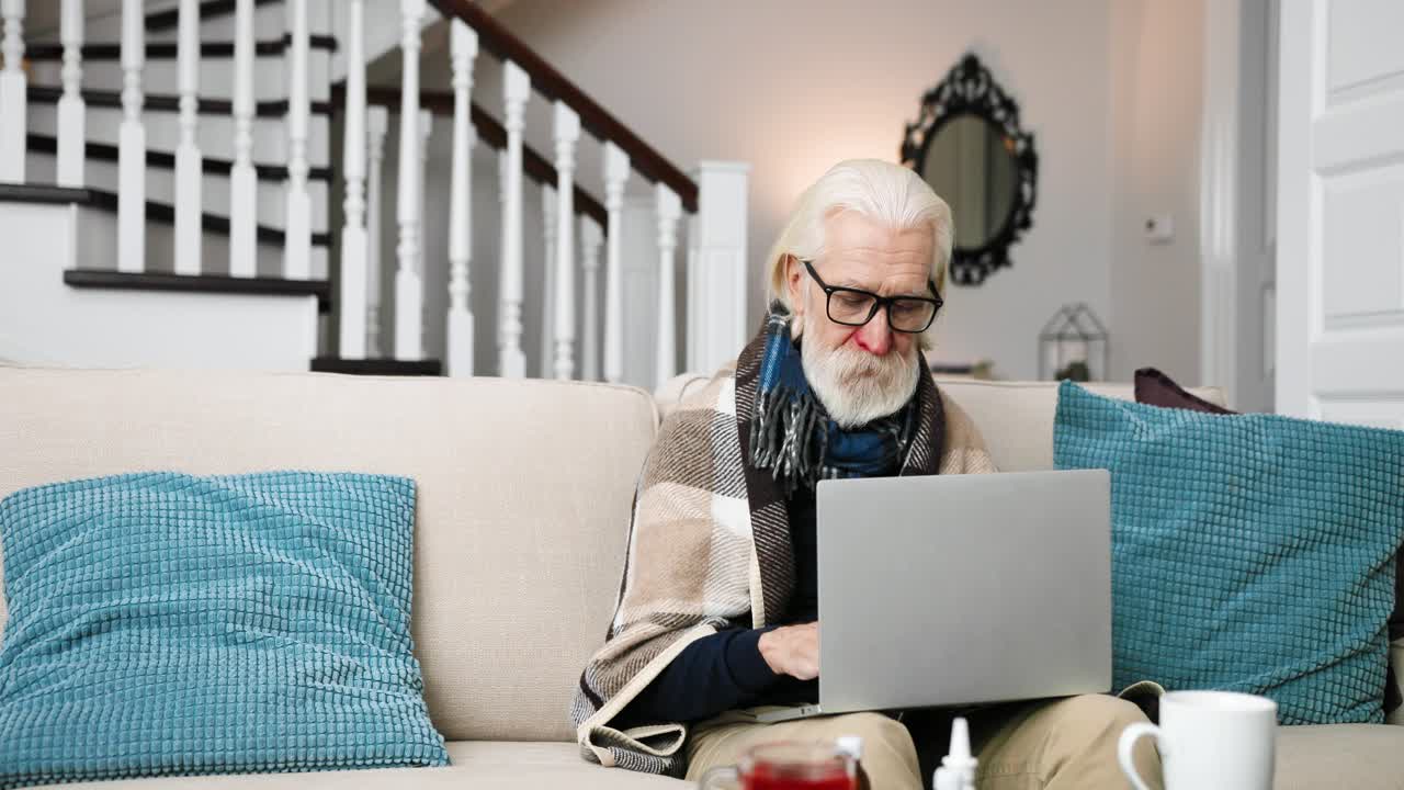 Sick old man in glasses sitting at home and typing on laptop