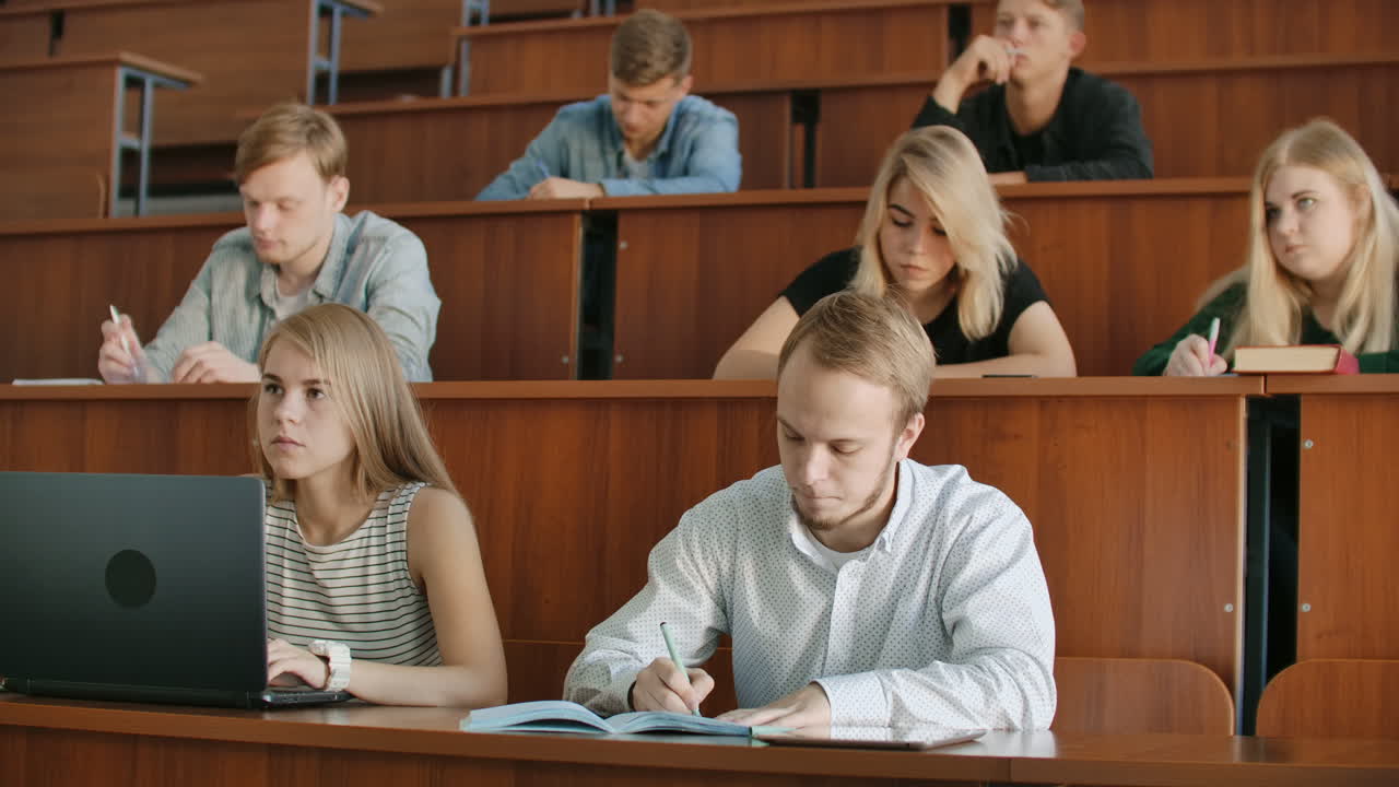 современные студенты, мужчины и женщины, сидят за столами в большом классе и пишут записки из лекций. получите образование для успешной карьеры.