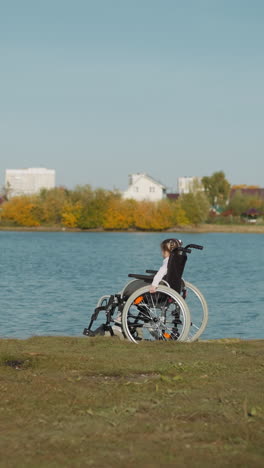 preschooler girl sits in wheelchair near river on sunny day. tranquil kid with long braids rests in summer. child with spinal cord injury on riverbank