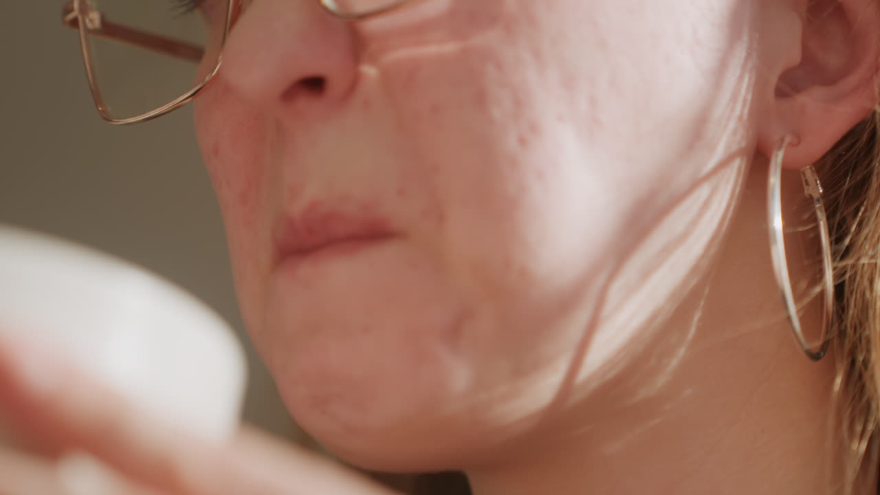 Close de mulher testando creme facial para pele acneica, usando óculos, experimentando do pote, expressão contemplativa, luz suave da manhã, momento íntimo de skincare mostrando poros e textura visíveis.