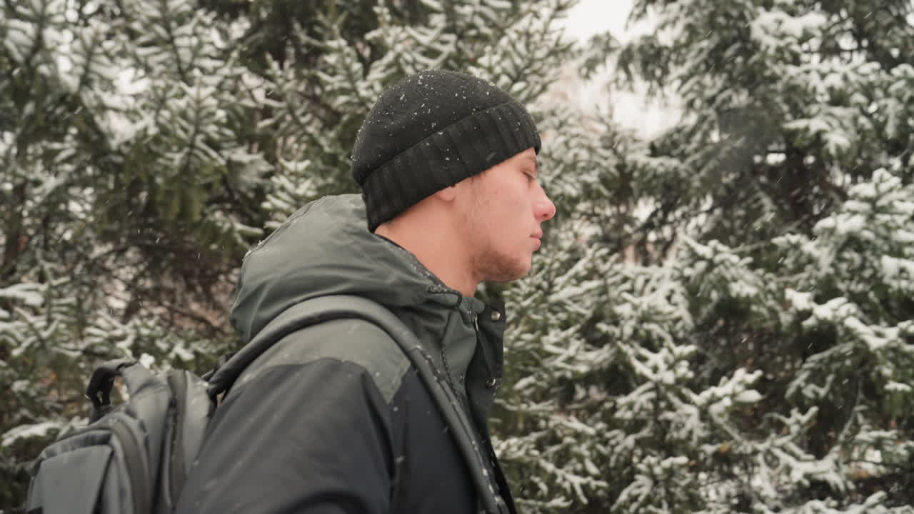 Man commuter walking snowy city park, purposeful profile with backpack and beanie, flurries drifting past residential buildings and evergreen trees, side view captures breath in cold air and steady
