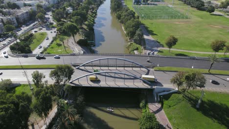 un puente urbano con autos cruzando un río con kayaks, amplios jardines y un campo de fútbol en un barrio residencial