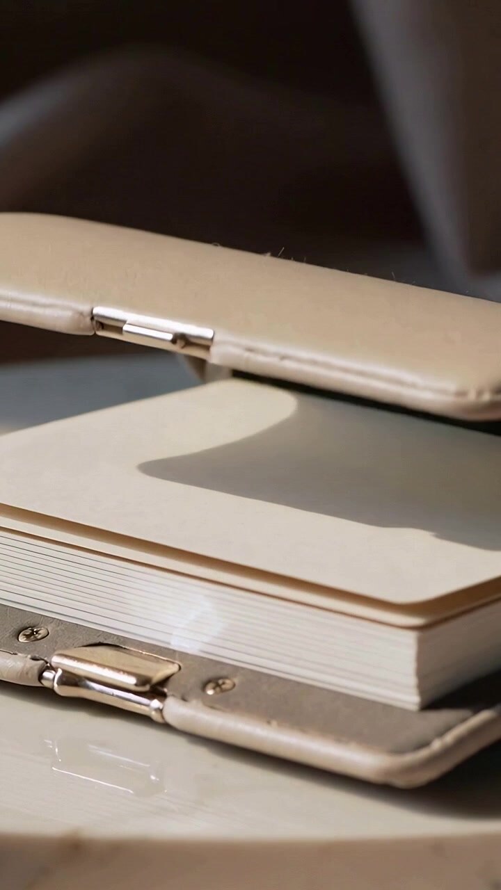 Vertical video: Lifting front, beige clutch opening on table, metal clasp showing cream note pages