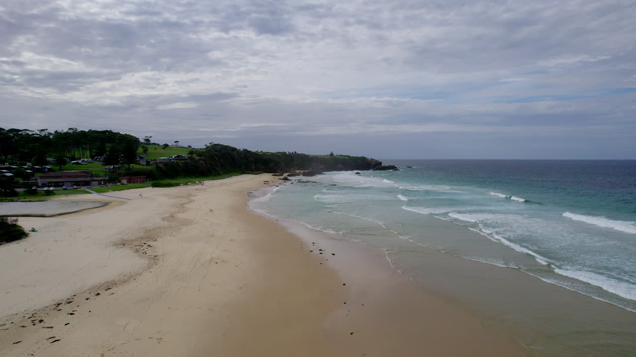 Ripresa cinematografica con drone che vola in avanti sopra Narooma Beach, mostrando acqua turchese, sabbia dorata, promontori rocciosi e una cittadina costiera sullo sfondo