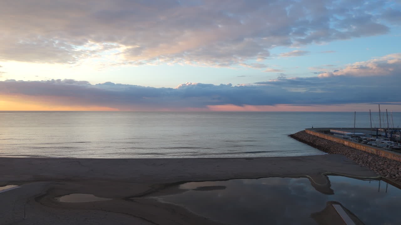 A stunning aerial lateral tracking shot at sunrise over Costa Dorada beach. The drone moves left, revealing the calm sea, golden sky, and marina, creating a serene and cinematic coastal landscape.