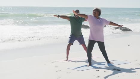 happy senior african american couple doing yoga and stretching at beach, in slow motion