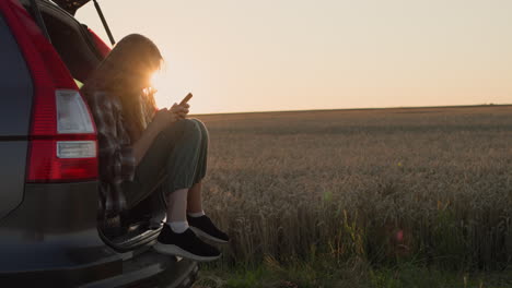 Child-sits-in-the-trunk-of-a-car,-uses-a-smartphone.-Against-the-backdrop-of-a-rural-landscape-where-the-sun-sets