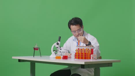 side view of asian man scientist looking at test tube then smiling and showing thumbs up gesture while working on the table with microscope in the green screen background laboratory