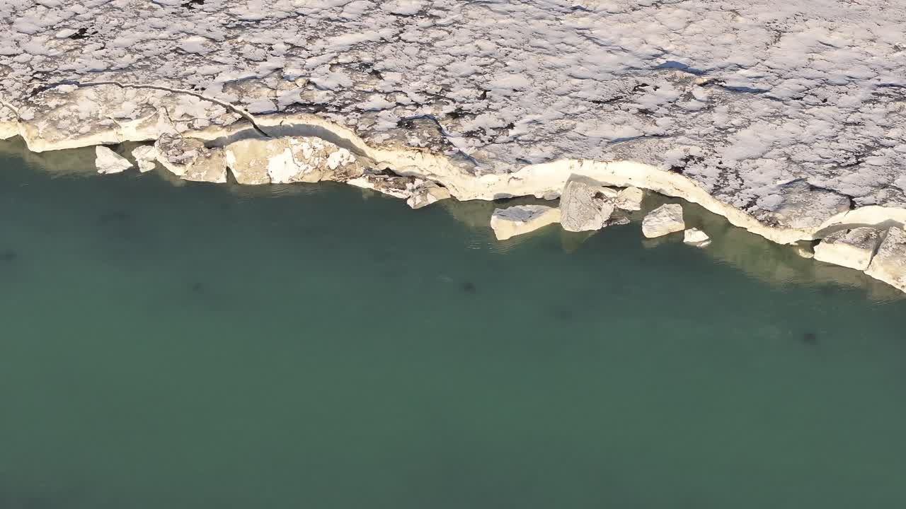 Aerial top-down view of the Urriðafoss waterfall in Iceland, where the icy river meets rugged, snow-covered terrain, showcasing the contrast between frozen and flowing water.