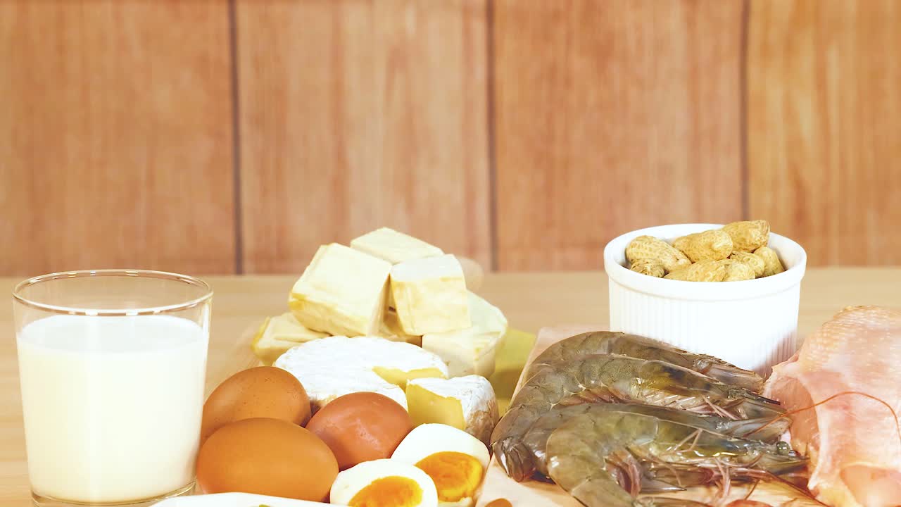 A variety of protein-rich foods including dairy, nuts, and meats are displayed on a wooden table in bright lighting