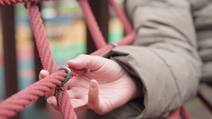 Child hand touching red rope net, winter coat sleeve, soft bokeh background, fingers exploring knot and weave, tactile curiosity captured in closeup, muted color palette, outdoor playground