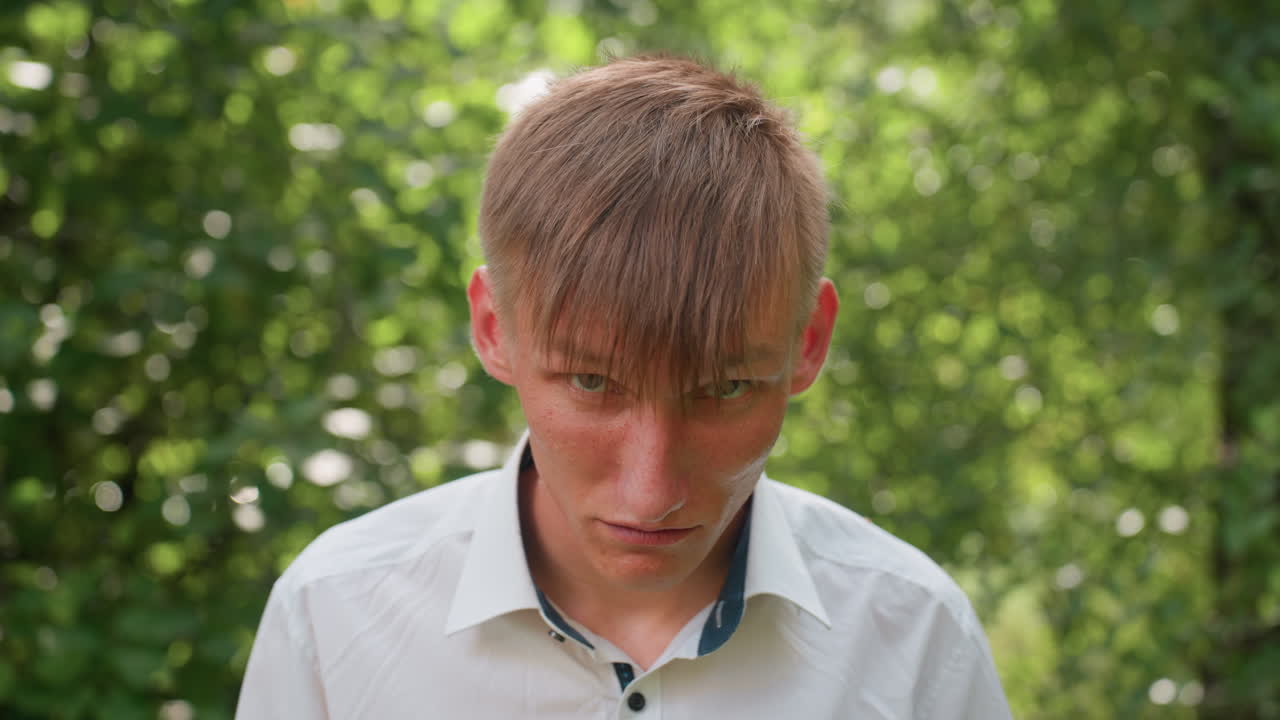 Young man in white shirt with glasses stands in forest under daylight, looking down with frowning face, expressing sadness and deep thought while surrounded by trees and green leaves