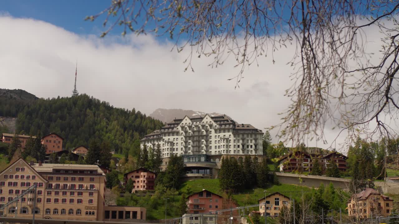 Großartige Architektur des Badrutt Palace Hotels, prominent am Hang in Sankt Moritz gelegen. Historisches Luxusresortgebäude, umgeben von Pinien unter blauem Himmel mit weißen Wolken.