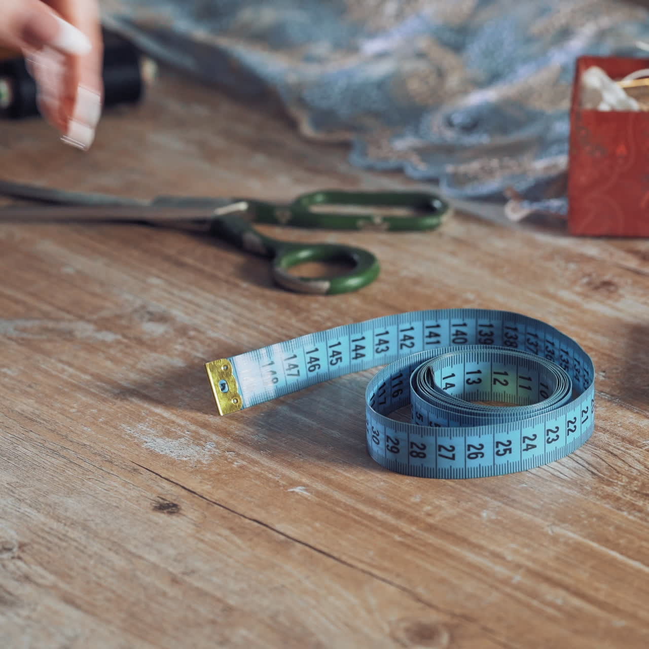 Seamstress is taking blue tape measure from the wooden table in atelier. Fingers of young female designer are taking the band with centimeter among sewing accessories.
