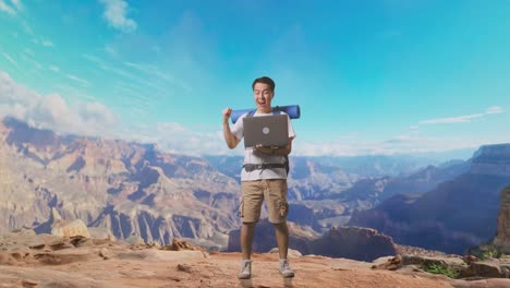 full body of asian male hiker with mountaineering backpack looking at a laptop and screaming goal celebrating the success while traveling at the top of mountain