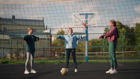 athletes standing on outdoor volleyball court, working out by turning their hands, volleyball rests in front, with sports facility and residential buildings in background