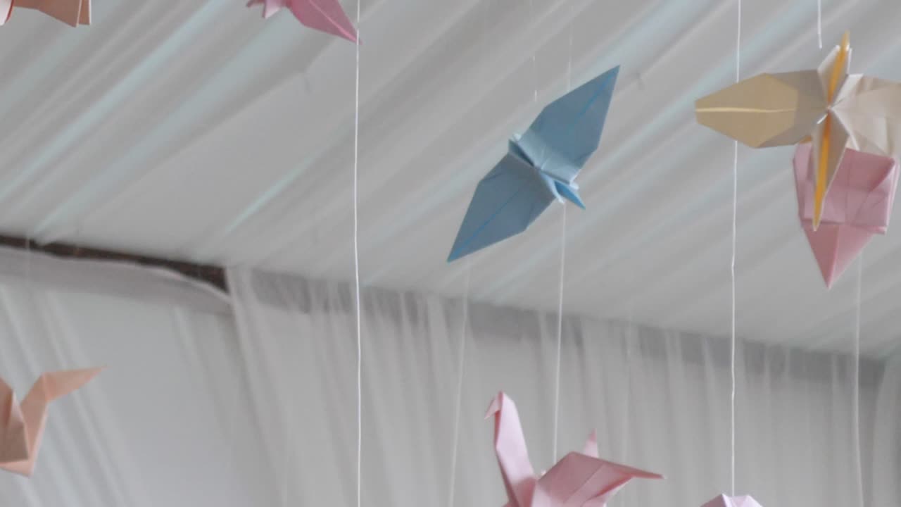 Close, slow motion footage of colourful pastel origami paper crane birds floating in breeze, hung from roof, blue, yellow, pink and orange