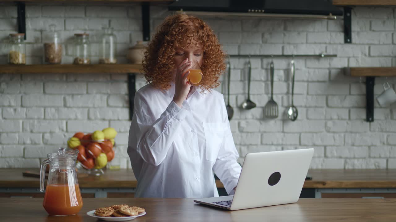 Redhead young woman with curly hair sit at table in cozy kitchen working at laptop computer, freelancer texting e-mail response to client, telework activity from home using internet and drinking juice