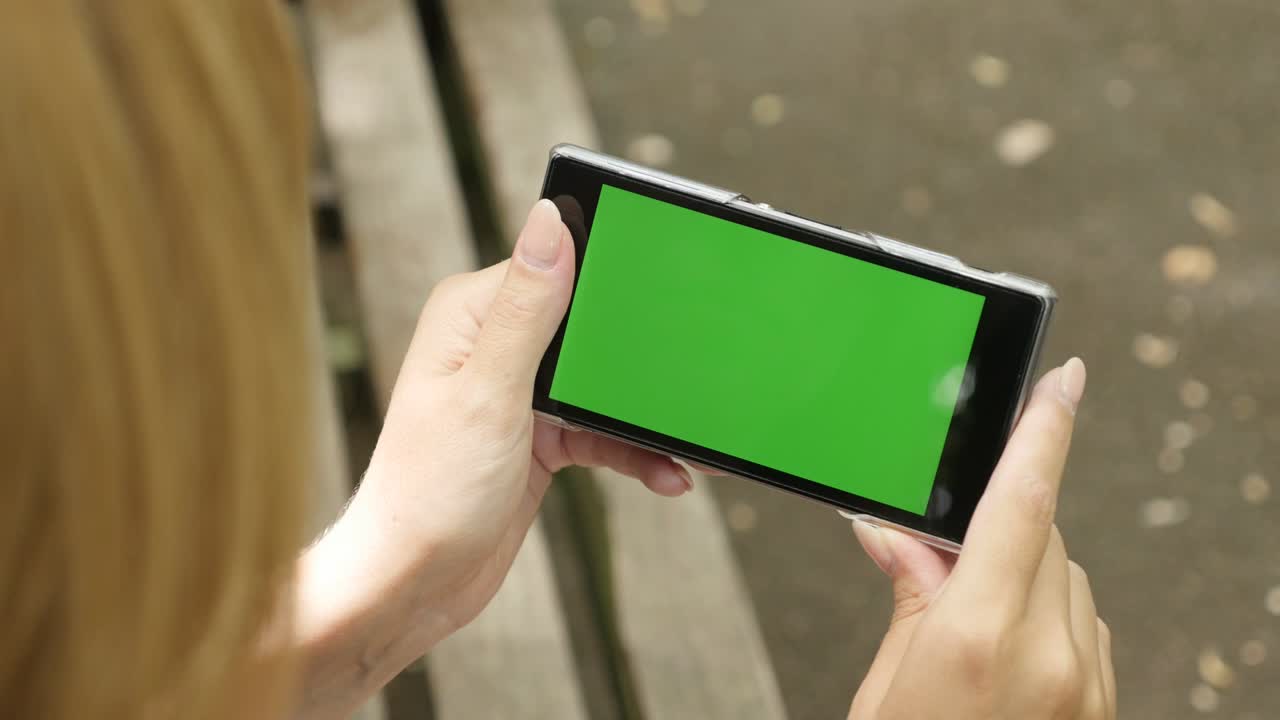 공원에서 금발의 여성이 벤치에서 휴식을 취하고 녹색 화면 전화를 들고 있습니다.