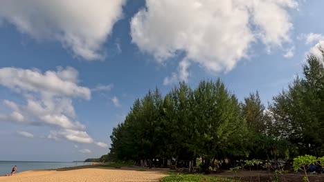 clouds move across a blue sky above a sandy beach and lush trees in phuket, thailand, creating a serene and tranquil scene