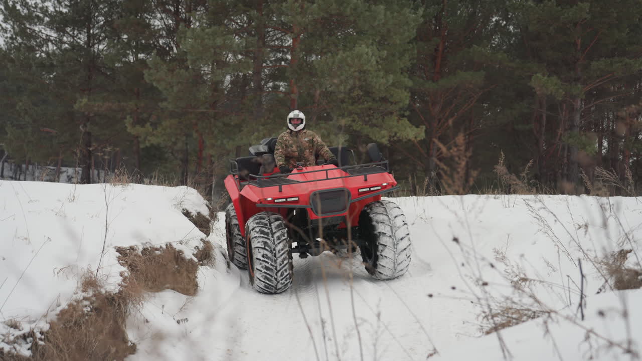 ATV rosso che scala un ripido pendio innevato, un pilota con casco in mimetica attacca la cresta. Le ruote smuovono neve e fango, un'angolazione ravvicinata della telecamera cattura gli spruzzi e il ruggito del motore. Atmosfera di intensa sfida off-road.