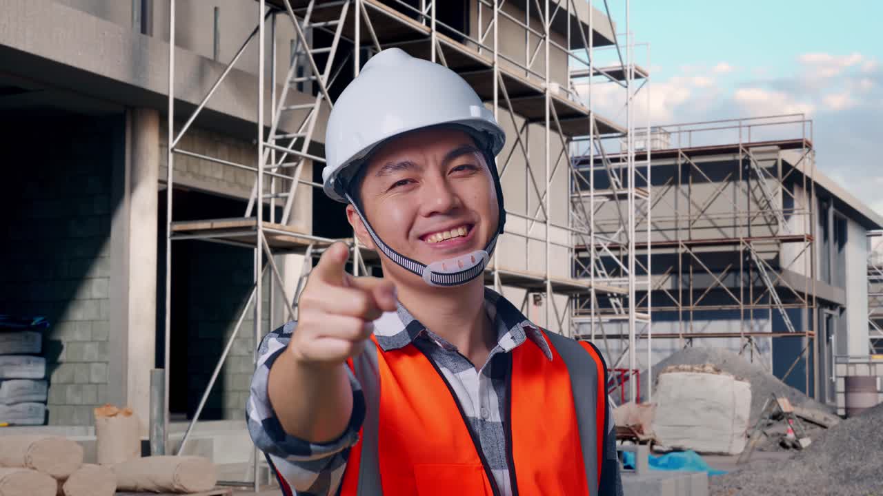 Close Up Of Asian Male Engineer With Safety Helmet Smiling And Touching His Chest Then Pointing At You While Standing At Building & Construction Site in Progress to New House