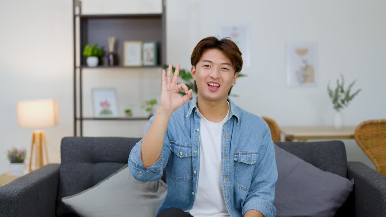 Un uomo asiatico sorridente con una camicia di jeans fa il segno di okay da un divano